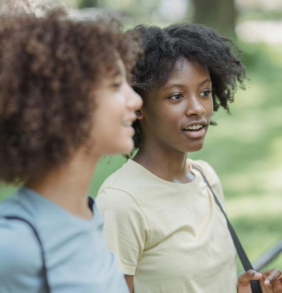 Two teenagers with curly hair chatting while walking outdoors in a park setting.